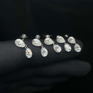 Puede incluir: Primer plano de varios pares de pendientes colgantes de diamantes. Cada pendiente presenta un peque&ntilde;o perno redondo y un diamante m&aacute;s grande en forma de pera suspendido debajo. Los diamantes brillan sobre un fondo oscuro, resaltando su claridad y brillo.