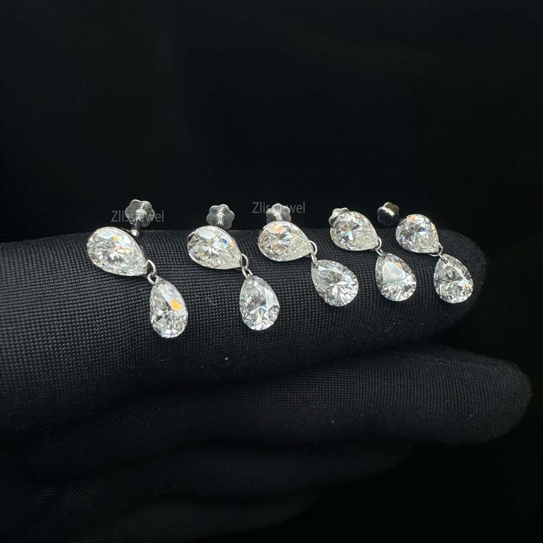 Puede incluir: Primer plano de varios pares de pendientes colgantes de diamantes. Cada pendiente presenta un peque&ntilde;o perno redondo y un diamante m&aacute;s grande en forma de pera suspendido debajo. Los diamantes brillan sobre un fondo oscuro, resaltando su claridad y brillo.