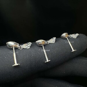 Puede incluir: Tres pendientes de bot&oacute;n plateados con detalles de piedras preciosas claras y facetadas. Cada pendiente presenta una piedra central redonda y una piedra triangular m&aacute;s peque&ntilde;a. Los pendientes se muestran sobre una superficie oscura y texturizada.