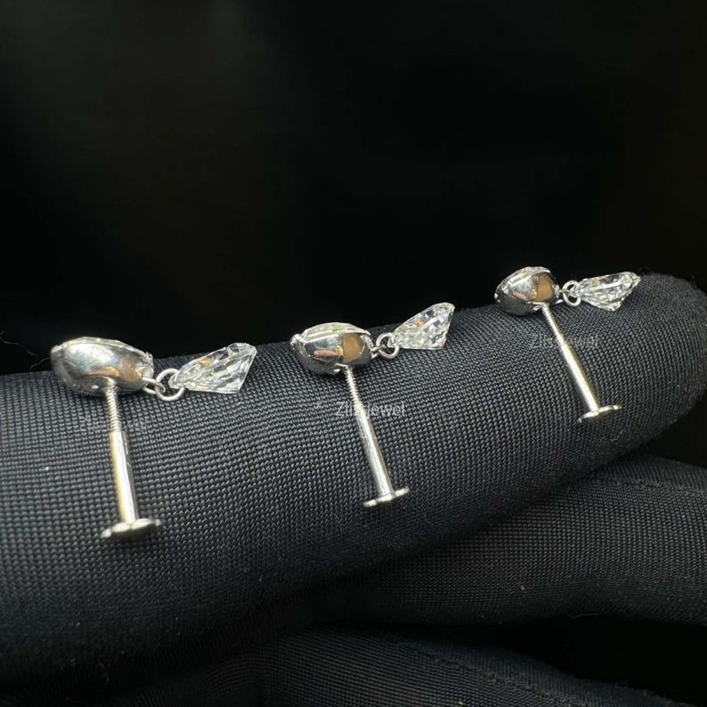 Puede incluir: Tres pendientes de bot&oacute;n plateados con detalles de piedras preciosas claras y facetadas. Cada pendiente presenta una piedra central redonda y una piedra triangular m&aacute;s peque&ntilde;a. Los pendientes se muestran sobre una superficie oscura y texturizada.