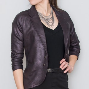 May include: A purple metallic blazer with a black v-neck shirt and a silver chain necklace. The blazer has a button closure and a pocket on the left side.