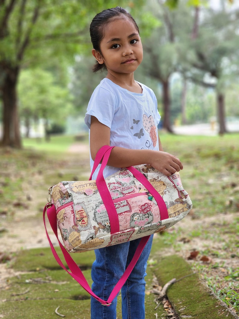 Day-To-Day Duffle Bag Sewing Pattern featuring the small size with a child model. The webbing is hot pink and the adjustable strap feature is shown.