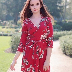 Puede incluir: Una mujer lleva un vestido rojo de envoltura con estampado floral. El vestido tiene un cintur&oacute;n de lazo y mangas tres cuartos. La mujer est&aacute; de pie en un camino en un parque.