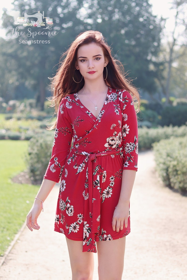 Puede incluir: Una mujer lleva un vestido rojo de envoltura con estampado floral. El vestido tiene un cintur&oacute;n de lazo y mangas tres cuartos. La mujer est&aacute; de pie en un camino en un parque.