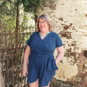 Puede incluir: Una mujer con un mono de mezclilla azul con un lazo en la cintura. Est&aacute; de pie frente a una pared de ladrillo con vegetaci&oacute;n verde.