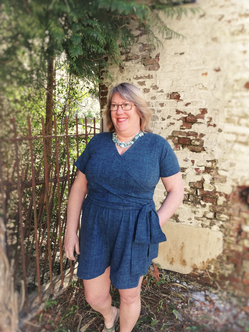Puede incluir: Una mujer con un mono de mezclilla azul con un lazo en la cintura. Est&aacute; de pie frente a una pared de ladrillo con vegetaci&oacute;n verde.