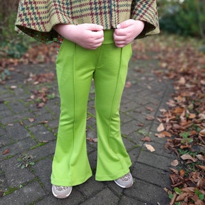 May include: A child wearing a green pair of flared pants with a brown and green plaid cape.