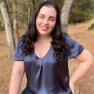 May include: A woman with long dark hair is wearing a dark blue satin top with a v-neck and short sleeves. She is smiling and looking at the camera. She is wearing a silver necklace with a small pendant.