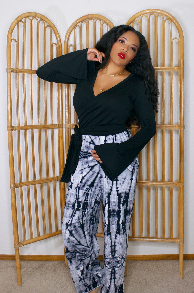 Puede incluir: Una mujer con un top negro de manga larga con cuello en V, mangas acampanadas y detalle de lazo, combinado con pantalones tie-dye en blanco y negro.