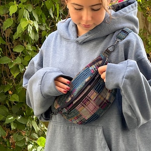 May include: A person wearing a light blue hoodie with a patchwork fabric fanny pack. The fanny pack has a black zipper and a silver buckle.