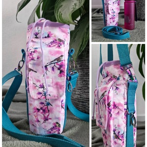 May include: A floral water bottle carrier with a teal strap. The carrier features a pink and white floral pattern with birds. The carrier has a small pocket and a drawstring closure. A pink water bottle is also visible.