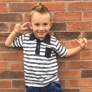 May include: A young boy with a spiky hairstyle wears a striped black and white polo shirt with a black pocket. The shirt has a black collar and blue buttons.