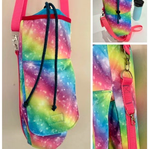 May include: A water bottle carrier with a vibrant galaxy print in shades of pink, blue, green, and yellow. It features a drawstring closure, a small front pocket, and a bright pink adjustable shoulder strap. The carrier is designed to hold a water bottle.