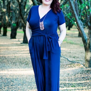 Puede incluir: Una mujer con un mono azul oscuro con una falda larga y fluida y un collar plateado. Est&aacute; de pie en un bosque de &aacute;rboles.