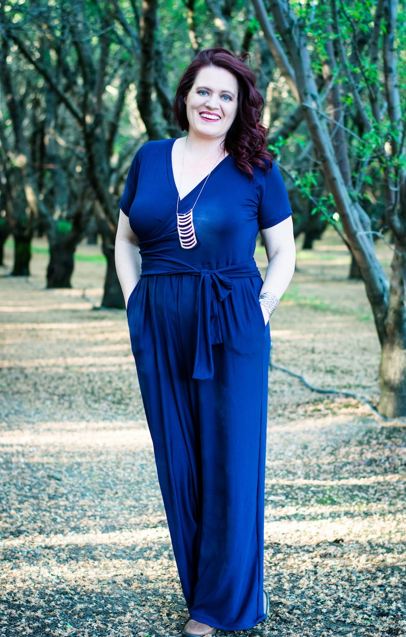 Puede incluir: Una mujer con un mono azul oscuro con una falda larga y fluida y un collar plateado. Est&aacute; de pie en un bosque de &aacute;rboles.