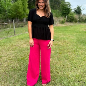 May include: A woman wearing a black short-sleeve top with a square neckline and a gathered waist, paired with bright pink wide-leg pants. The outfit is completed with sandals. The background features green grass and trees.