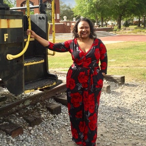 Puede incluir: Una mujer con un mono floral negro y rojo con un corpi&ntilde;o de estilo envolvente. Est&aacute; de pie frente a una v&iacute;a de tren con una barandilla amarilla.