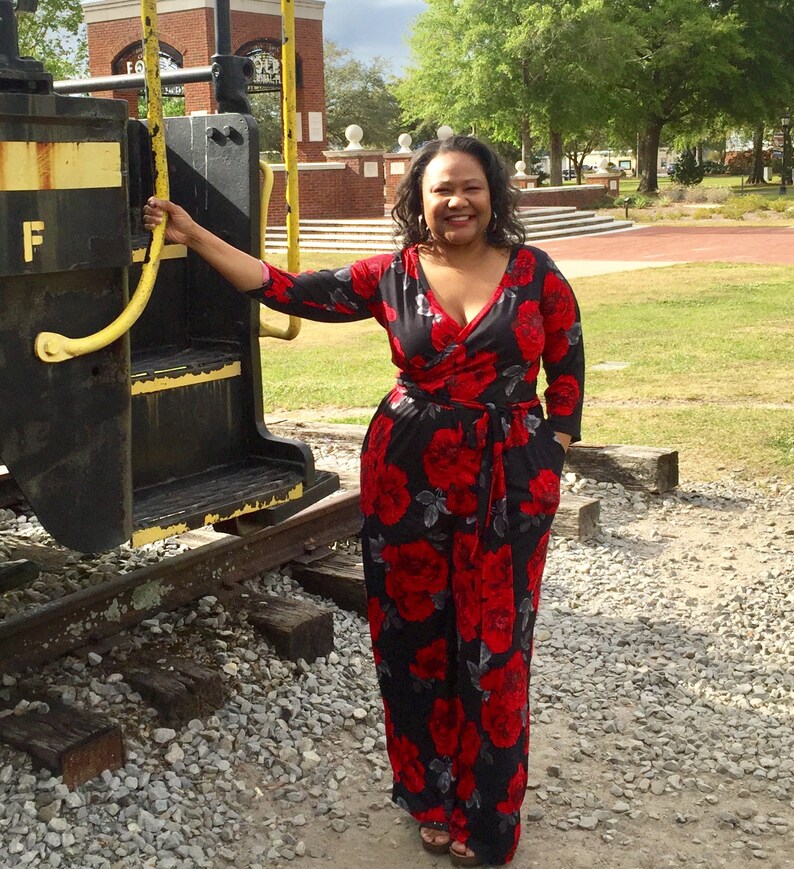Puede incluir: Una mujer con un mono floral negro y rojo con un corpi&ntilde;o de estilo envolvente. Est&aacute; de pie frente a una v&iacute;a de tren con una barandilla amarilla.