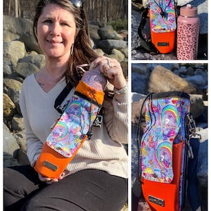 May include: A water bottle carrier with a colorful, whimsical design featuring unicorns, rainbows, and psychedelic patterns. The carrier has an orange base and a black adjustable strap. A matching water bottle with a pink leopard print is also visible.