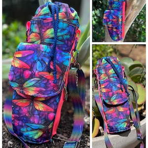May include: A colorful crossbody bag with a dragonfly pattern in shades of purple, blue, pink, and green. The bag has a drawstring closure, a small front pocket, and a rainbow-colored strap. The interior is lined with a bright pink fabric.