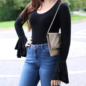 May include: A woman wearing a black long-sleeved top with ruffled cuffs and blue jeans. She is carrying a light brown chain strap clutch.