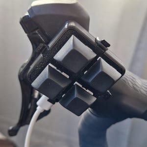 Puede incluir: Primer plano de un dispositivo negro con cuatro botones grises en forma de pirámide sujetos a un manillar de bicicleta. Un cable blanco está conectado al dispositivo. El manillar y el dispositivo son negros.