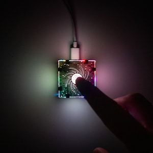 Könnte beinhalten: Ein kleines, quadratisches elektronisches Gerät mit einem regenbogenfarbenen Rand und einem weißen Knopf in der Mitte. Der Knopf leuchtet und ein Finger drückt darauf.