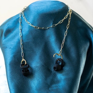 May include: A gold-colored chain necklace with two dangling black earbud pendants. The earbuds are attached to the chain with gold-colored clasps. The necklace is displayed on a teal-colored fabric.