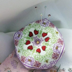 May include: A white pincushion with a floral design and red strawberries. The pincushion is surrounded by decorative pins, scissors, and beads on a pink floral fabric.