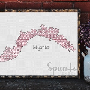May include: A cross-stitch pattern of the Liguria region of Italy, with a pink and white geometric design. The pattern is framed and the text "Liguria" is written below the pattern.