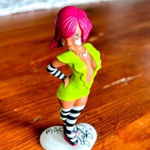 May include: A figurine of a woman with pink hair, wearing a neon green dress, black and white striped thigh-high socks, and armbands. The figurine stands on a white heart-shaped base with the word "MAC" and other text. The figurine has heart-shaped face paint.