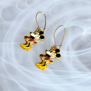 May include: A pair of gold-coloured dangle earrings featuring a cartoon mouse character with black ears, a white face, red shorts, and yellow shoes. The earrings have a hook closure and are set against a white and grey background.