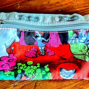 May include: A small, rectangular zippered pouch with a colorful graphic print. The print features a cartoon-like illustration with figures in a vibrant red, pink, and green landscape. The top edge has the brand name "LESORTSAC" printed on it.