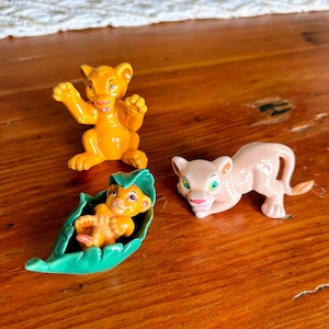 May include: Three small, colorful figurines of lion cubs. One is orange and posed with its arms up. Another is a light pink lioness. The third is a cub in a green leaf.