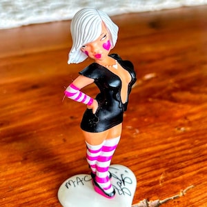 May include: Figurine of a woman with white hair, pink heart-shaped cheek accents, and pink lips. She wears a black dress, pink and white striped armbands and socks, and pink shoes. The figurine stands on a white heart-shaped base with text.