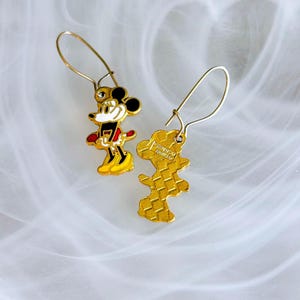 May include: A pair of gold-coloured Mickey Mouse drop earrings. Each earring features a detailed enamel design of Mickey Mouse in his classic outfit, with black, red, yellow, and white colours. The earrings have a hook closure.