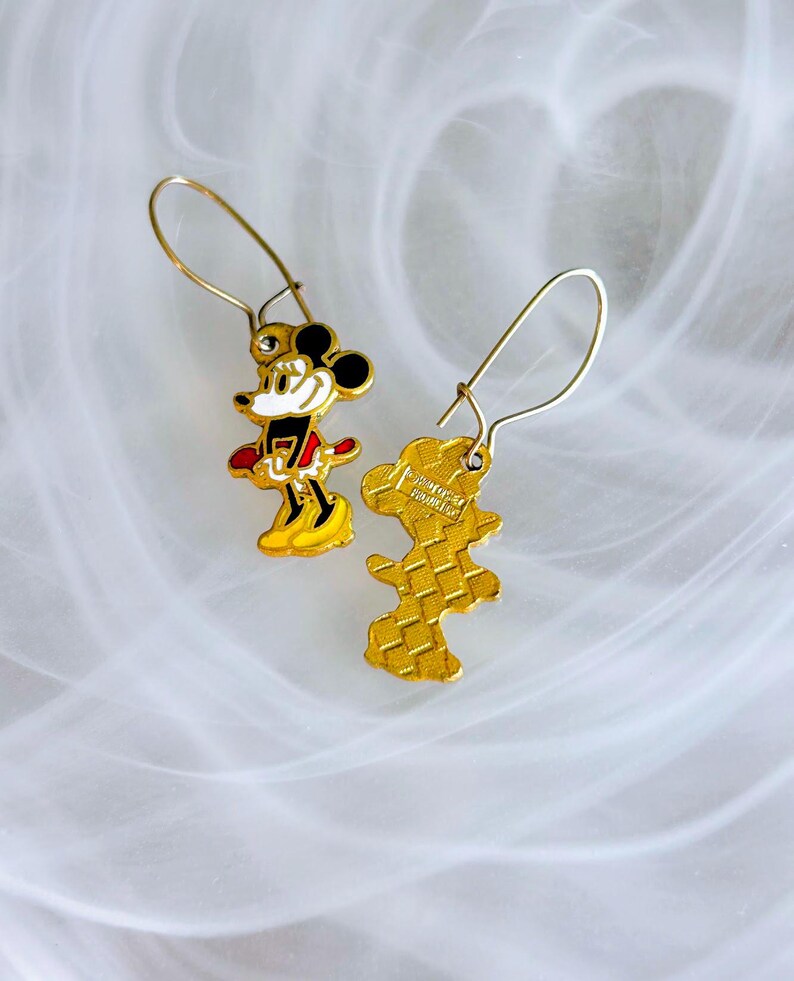May include: A pair of gold-coloured Mickey Mouse drop earrings. Each earring features a detailed enamel design of Mickey Mouse in his classic outfit, with black, red, yellow, and white colours. The earrings have a hook closure.