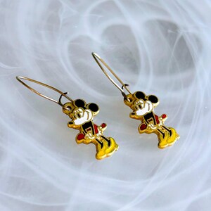 May include: A pair of gold-coloured Mickey Mouse dangle earrings. Each earring features a miniature Mickey Mouse figure with black ears, a white face, red shorts, and yellow shoes. The earrings have a simple hook design.
