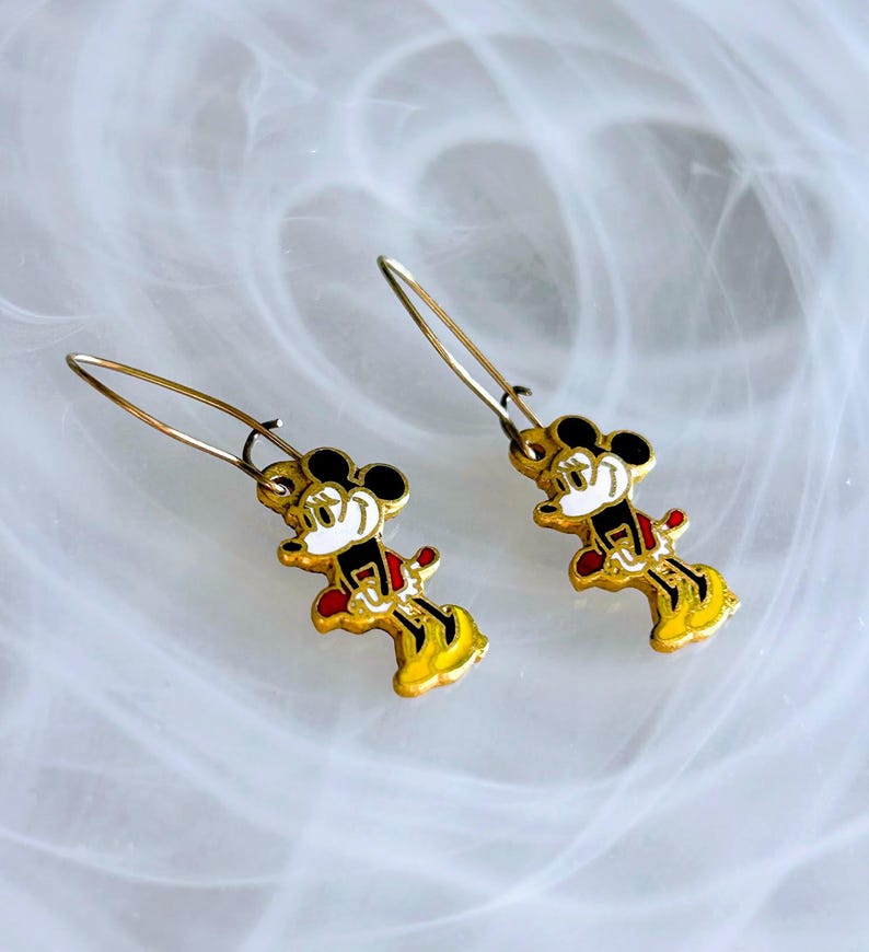 May include: A pair of gold-coloured Mickey Mouse dangle earrings. Each earring features a miniature Mickey Mouse figure with black ears, a white face, red shorts, and yellow shoes. The earrings have a simple hook design.