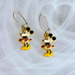 May include: A pair of gold-coloured Mickey Mouse dangle earrings. Each earring features a colourful enamel design of Mickey Mouse in his classic outfit, with black ears, a red shirt, and yellow shoes. The earrings have a simple hook closure.