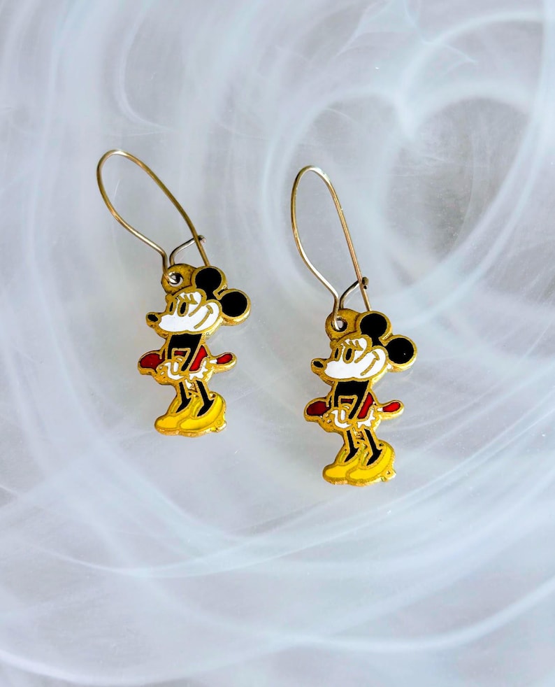 May include: A pair of gold-coloured Mickey Mouse dangle earrings. Each earring features a colourful enamel design of Mickey Mouse in his classic outfit, with black ears, a red shirt, and yellow shoes. The earrings have a simple hook closure.