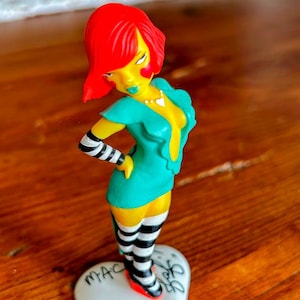 May include: A figurine of a woman with red hair, yellow skin, and a teal dress. She wears black and white striped socks and armbands. The figurine stands on a white heart-shaped base with the word "MAC" and other text written on it.