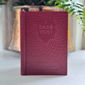 May include: A burgundy-colored book with the title "I DARE YOU!" embossed on the cover. The cover features a textured pattern and a silhouette. The spine reads "I DARE YOU!" and "DANFORTH".