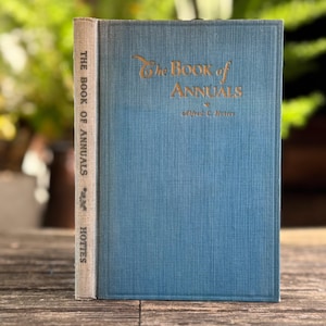 May include: A vintage book titled "The Book of Annuals" with a blue cover and gold lettering. The spine is a light beige color with the title and author's name. The book is standing on a wooden surface.