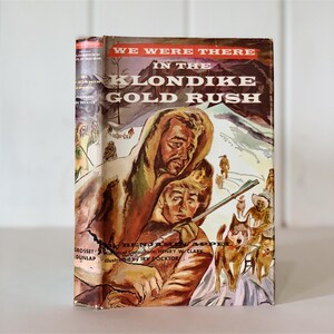 May include: A vintage book cover with a colorful illustration of two men in winter clothing with a dog sled team in the background. The title of the book is "We Were There in the Klondike Gold Rush".