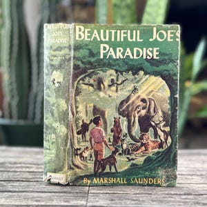 May include: Vintage hardcover book titled "Beautiful Joe's Paradise" by Marshall Saunders. The cover features a forest scene with an elephant, boy, and various animals. The book has a green cover with white text.