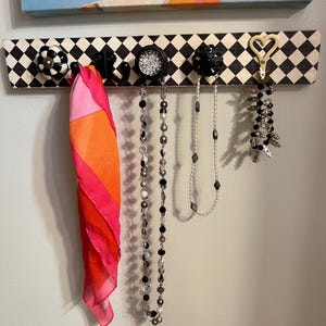 Può includere: Un organizer da parete a scacchi bianchi e neri con pomelli e ganci decorativi. Una sciarpa rosa e arancione è appesa a un gancio, accanto a diverse collane. L'organizer è montato su una parete azzurra.