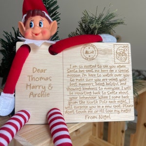 May include: A wooden elf postcard with a red and white striped elf holding the postcard. The postcard has a message from Nigel the elf to Thomas, Harry, and Archie. The message says, "I am so excited to see you again. Santa has sent me here on a special mission. I'm here to watch over you to make sure you are using your best manners, being helpful and showing kindness to everyone. I will be reporting back to Santa about your behaviour. When I come back from the North Pole each night, I will surprise you in a new spot. Please don't touch me or I'll lose my magic! From Nigel."