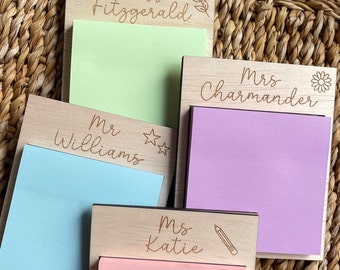 May include: Wooden sticky note holders with engraved names and decorative elements. Each holder contains a pad of colorful sticky notes. The names "Miss Fitzgerald", "Mr Williams", "Mrs Charmander", and "Ms Katie" are visible.