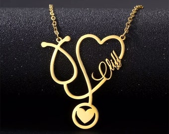May include: Gold-colored necklace featuring a stethoscope design with a heart-shaped pendant. The pendant includes the name "Elly" in a cursive font. The necklace is on a dark background.